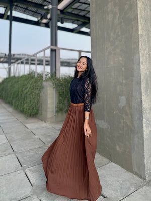 Woman in black top and brown pleated skirt standing outdoors near a concrete wall.