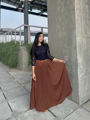 Woman wearing a black top and brown pleated skirt standing outdoors near a concrete wall.