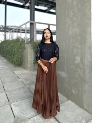 Woman wearing a black top with lace details and a brown pleated skirt standing outdoors.
