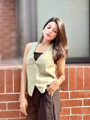 Woman wearing a light green vest over a black top, standing against a brick wall.