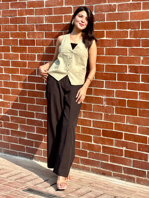 Woman in a sleeveless vest and dark pants standing against a brick wall.