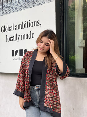 Woman wearing a patterned short kimonos and jeans leaning against a wall with a sign in the background.