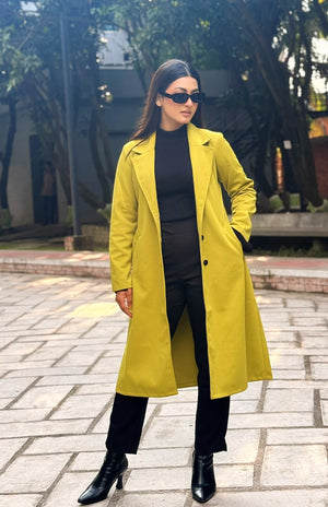 Woman wearing a yellow coat and black outfit standing outdoors on a paved area with trees in the background.