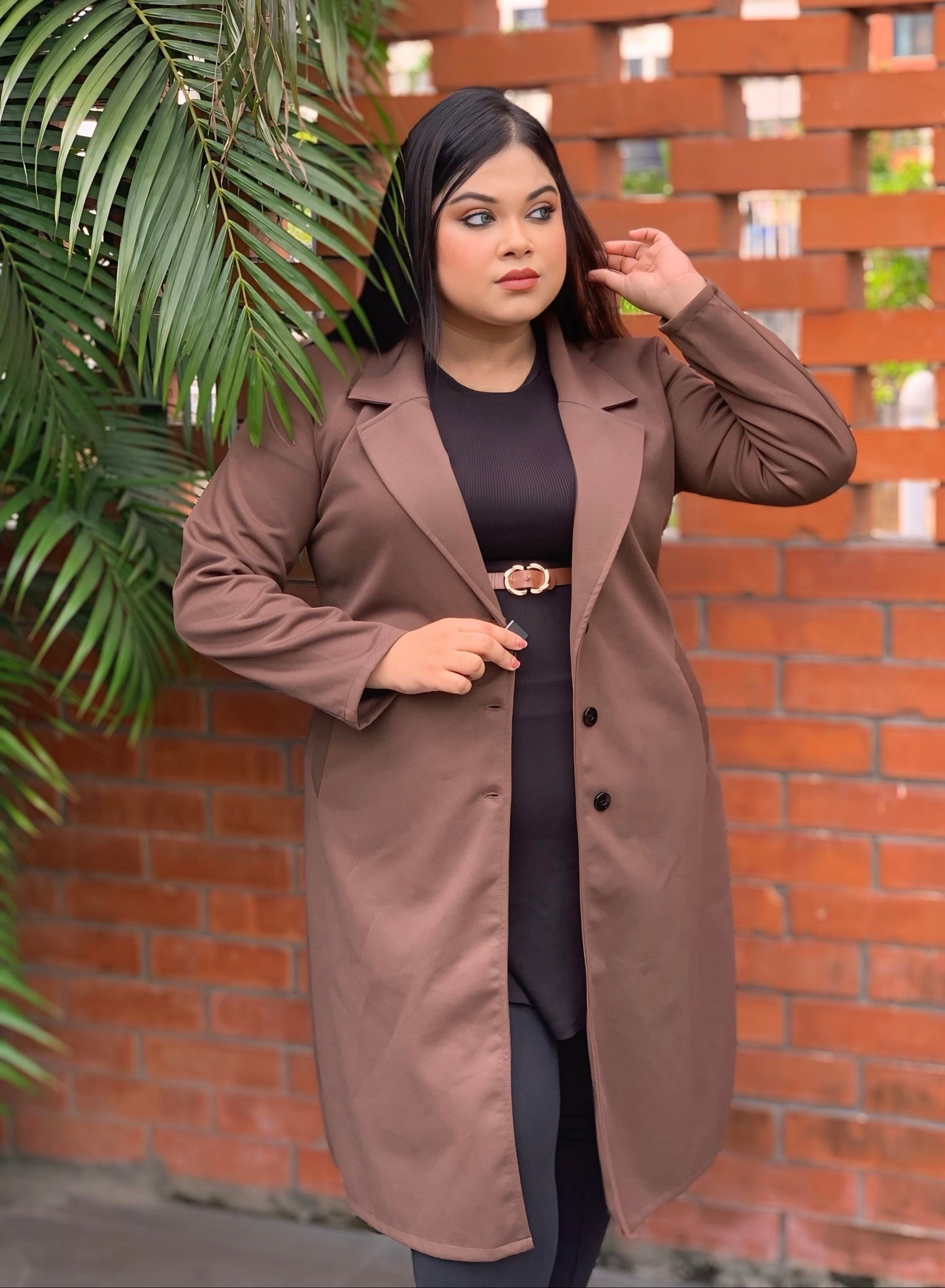 Woman wearing a brown coat standing against a brick wall with plants.