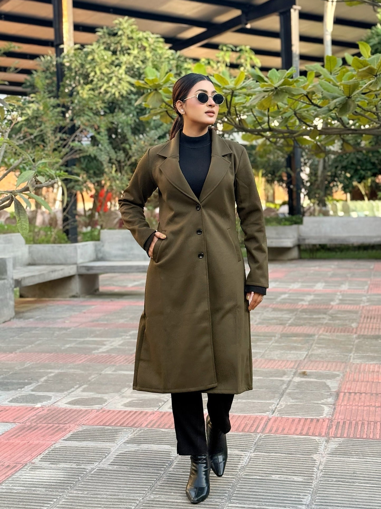 Person wearing a long olive green coat in an outdoor setting with trees and benches.