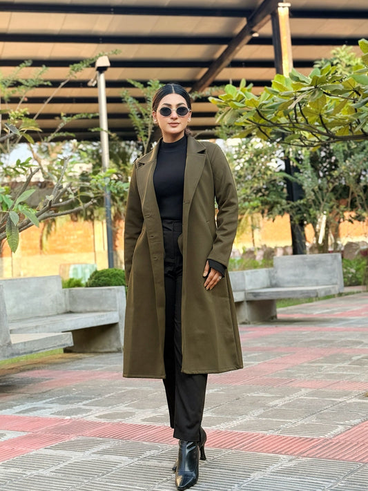 Woman in a green coat standing outdoors on a paved area with trees and benches in the background.