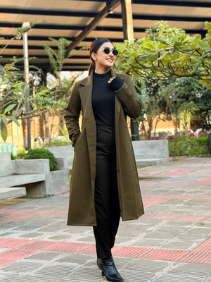 Woman in a long green coat standing outdoors with trees and a building in the background