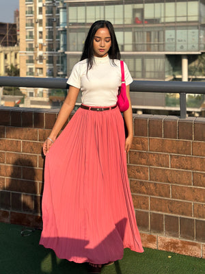 Woman wearing a white top and pink pleated skirt standing on a rooftop with buildings in the background.