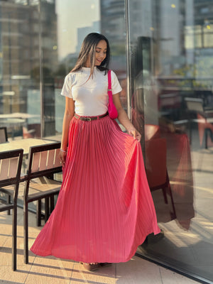 Woman in a white top and pink pleated skirt standing in an outdoor setting with chairs and tables.