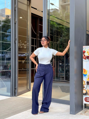 A woman wearing a white short-sleeve mock neck top and navy blue wide-leg trousers stands in front of a modern glass building entrance.