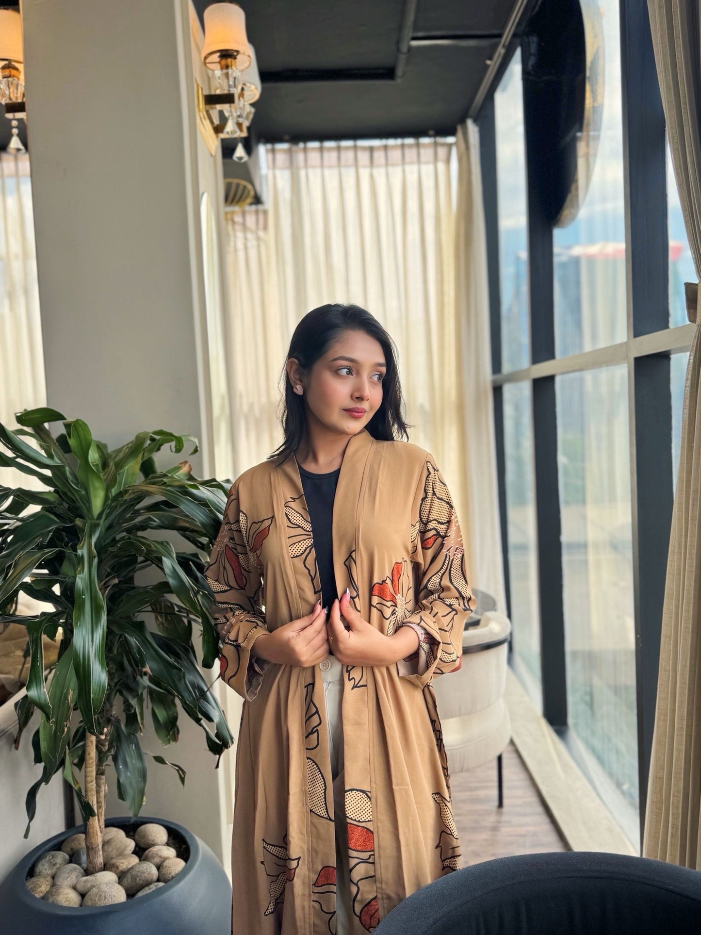 Woman in a beige robe with floral patterns standing in a modern room with large windows.