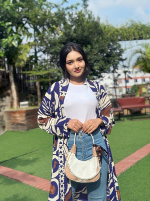 Woman wearing a long patterned shrug-style jacket and jeans with a handbag outdoors.