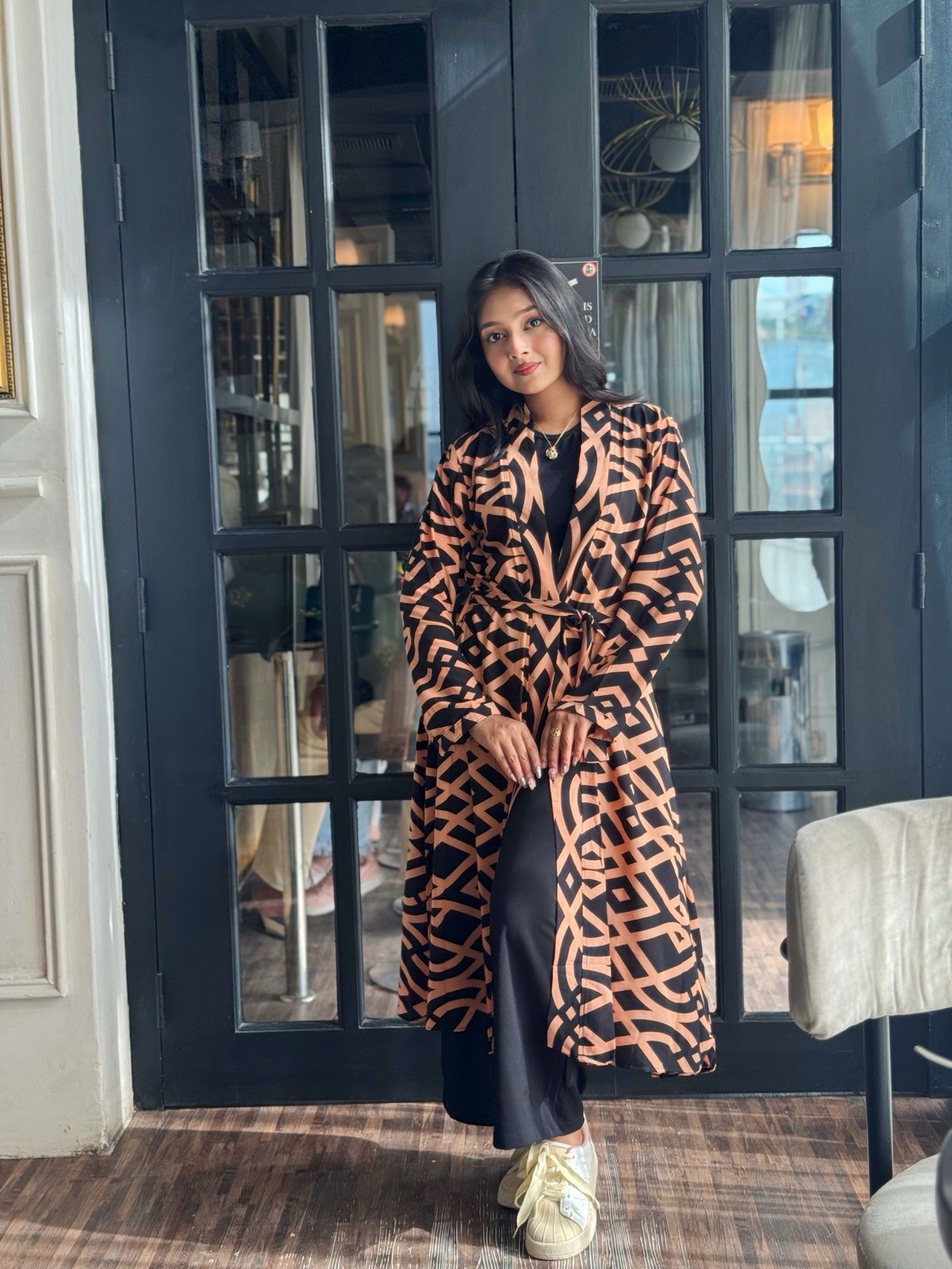 Woman wearing a Woman wearing a long shrug outerwear in black and orange standing in front of a glass door.