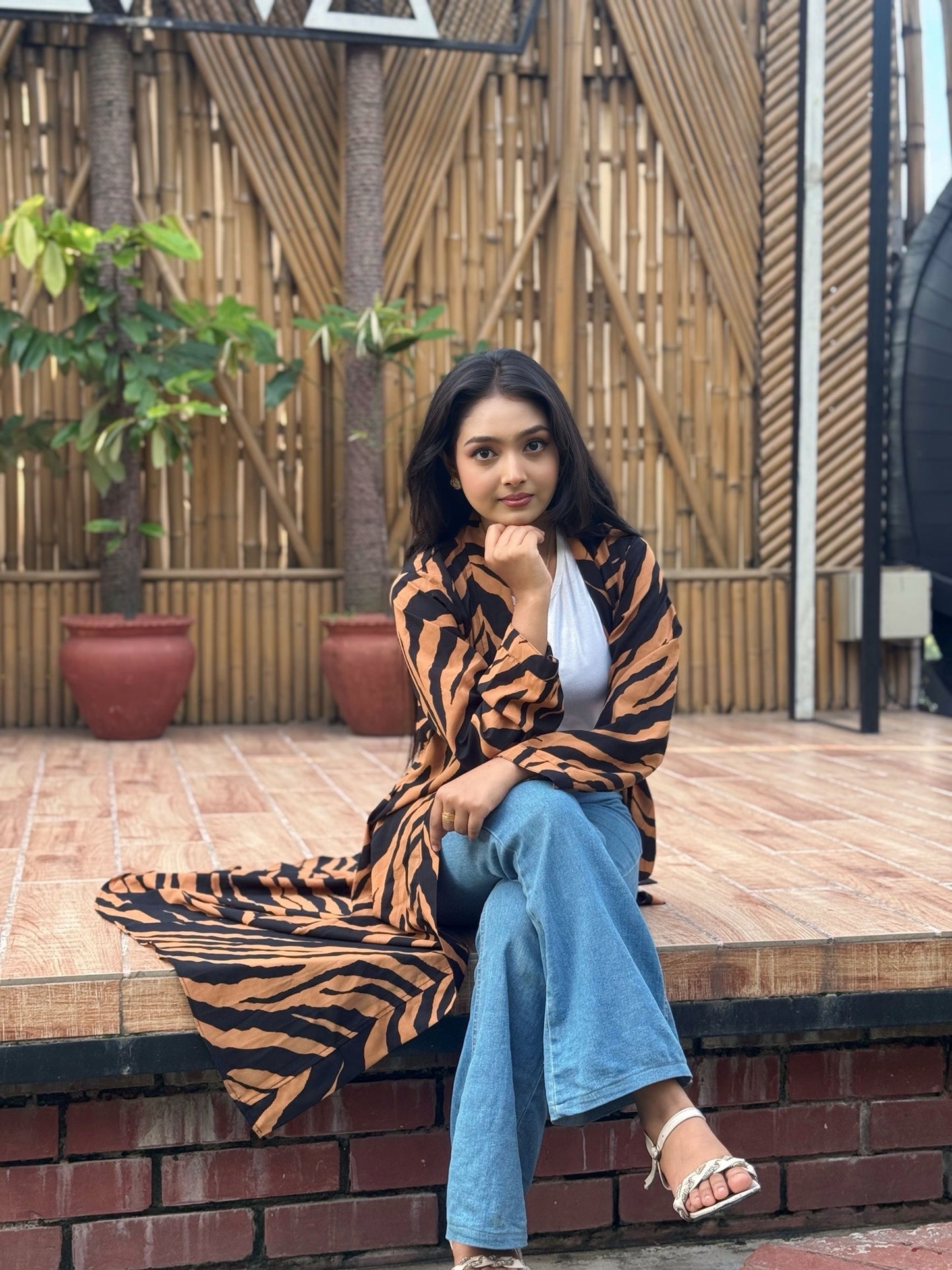 Woman sitting on a ledge wearing a tiger-striped long shrug and blue jeans, with a bamboo wall background.