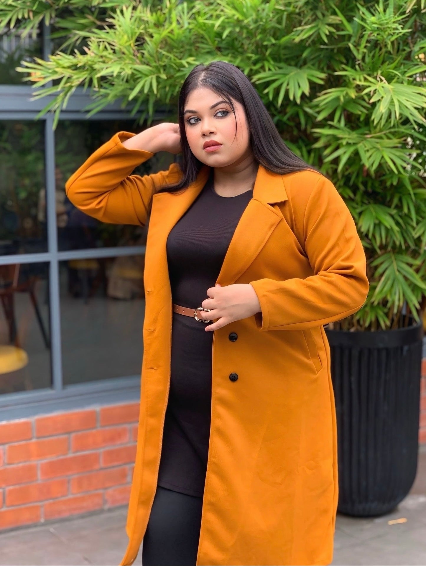 Woman wearing a mustard yellow coat standing outdoors with greenery in the background