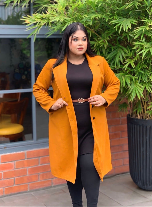 Woman wearing a mustard yellow coat standing outdoors with greenery and a building in the background