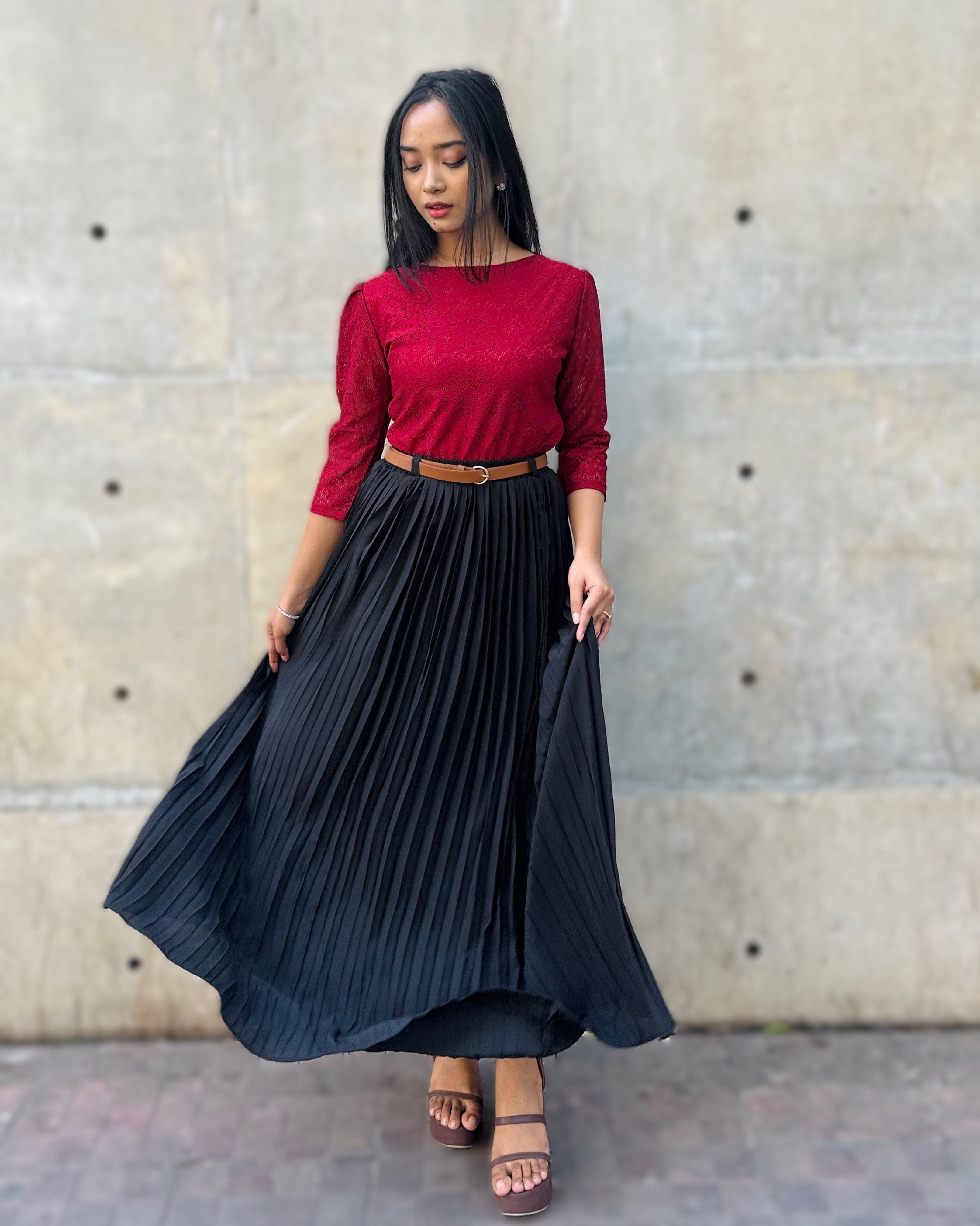 Woman wearing a red top and black pleated skirt against a concrete wall.