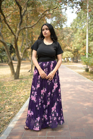 Woman in a black top and purple floral dress standing outdoors with trees and grass in the background TrendyBuzz