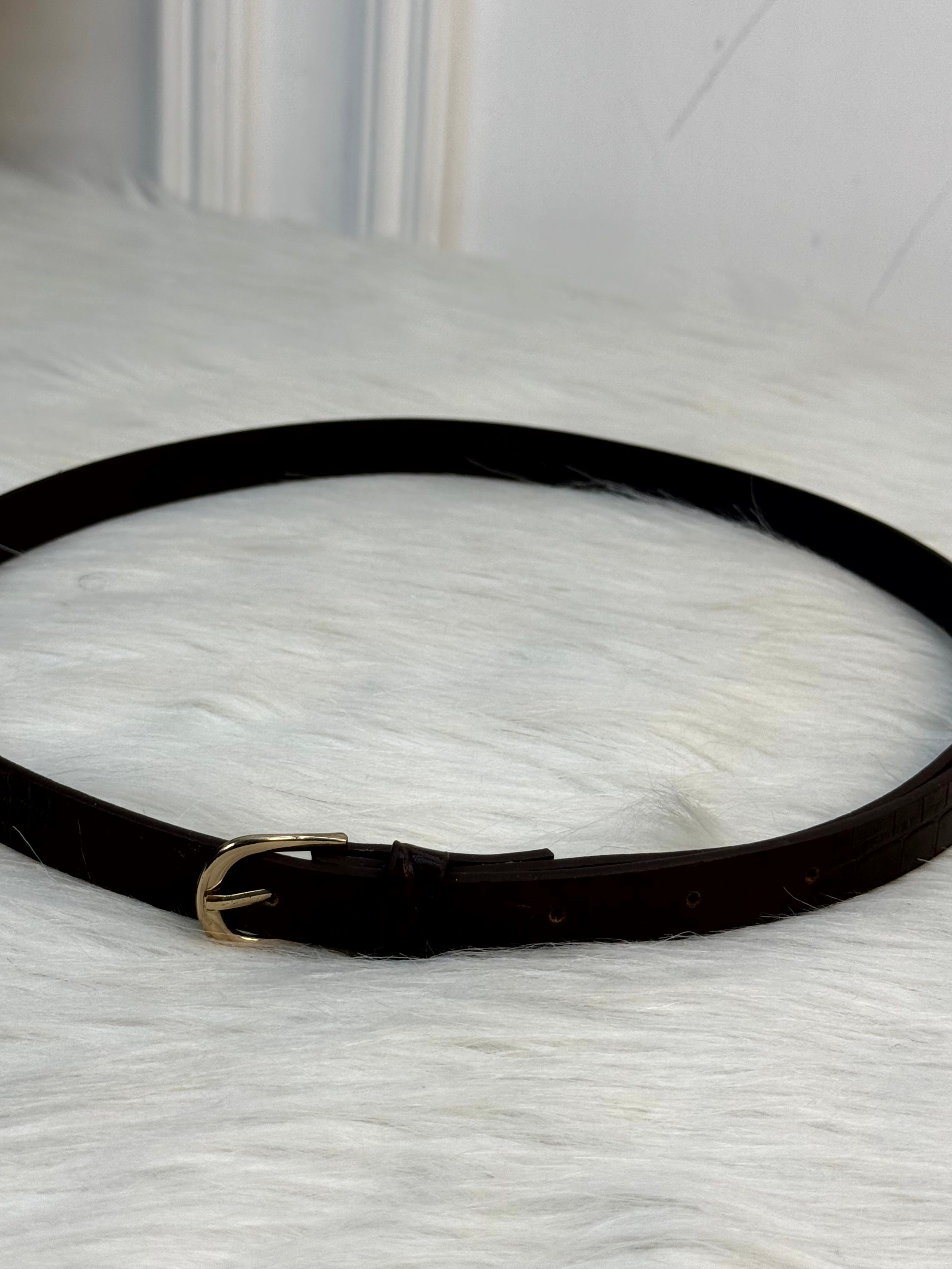 A close-up of a dark brown leather belt with a crocodile-embossed texture and a polished gold-tone buckle, resting on a white faux fur surface. rendy Buzz BD.