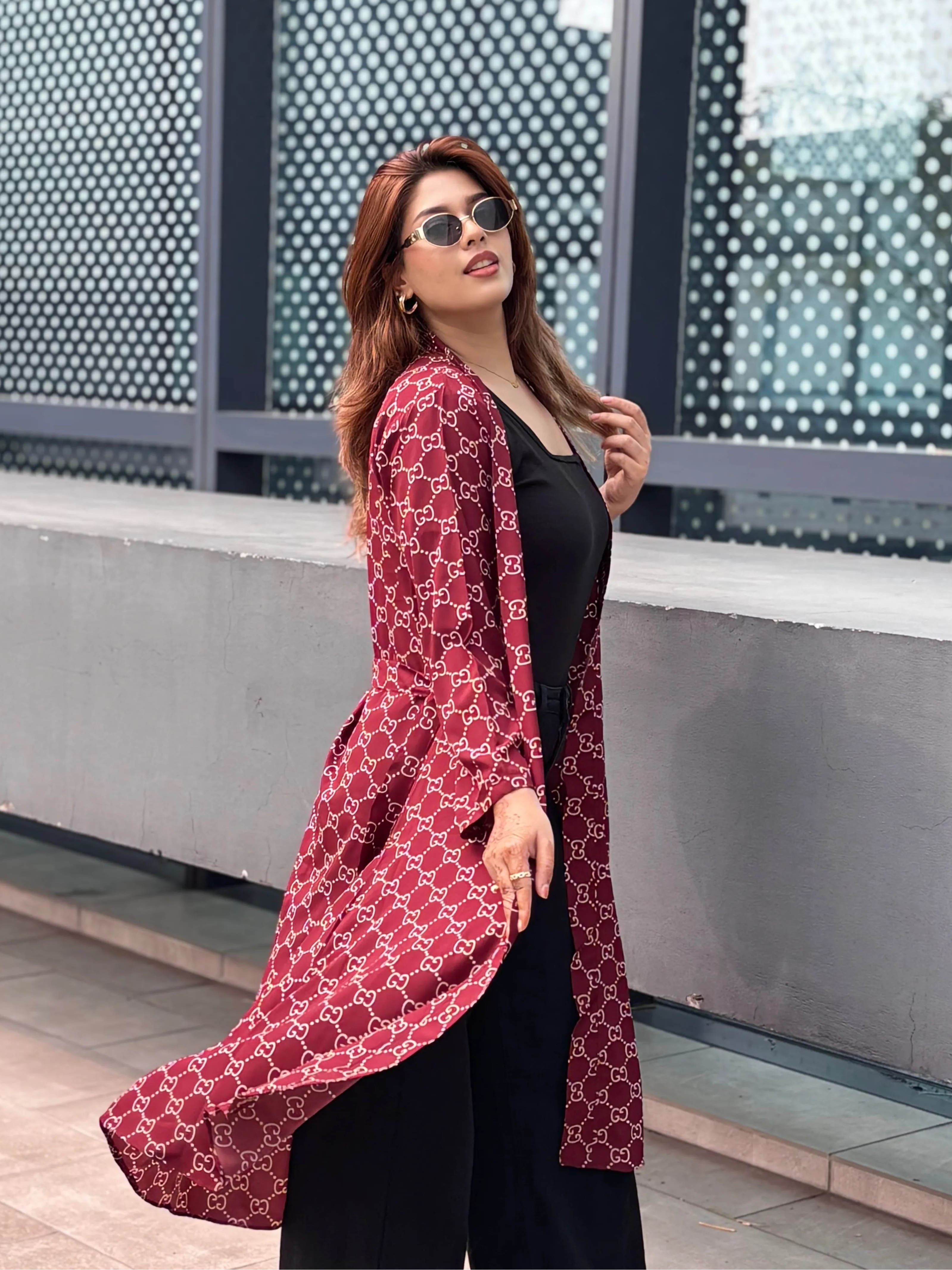 Woman wearing a red patterned long Shrug over a black outfit, standing in front of a modern building.