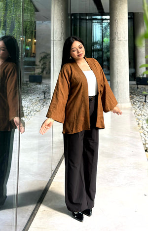 A woman modeling a brown open-front kimono with wide sleeves over a white top and black wide-leg trousers, standing in a modern walkway with glass walls and columns.