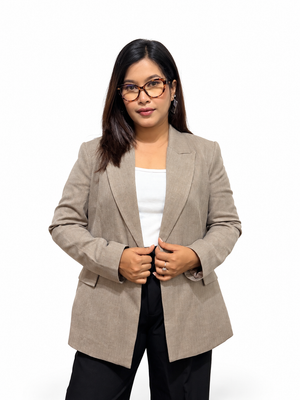 A woman wearing a light taupe-beige blazer over a white top and black pants, standing against a plain white studio background.