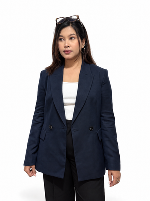 A woman modeling a navy blue double-breasted blazer over a white top and black trousers, standing against a plain white studio background with sunglasses on her head.
