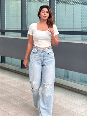 A woman walking outdoors wearing a white short-sleeve crop top and light-wash high-waisted wide-leg jeans with distressed details. TREND BUZZ