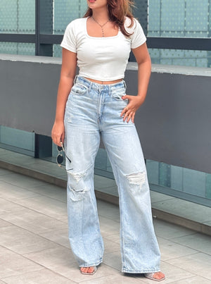A woman modeling a white short-sleeve crop top and light-wash wide-leg distressed jeans, standing outdoors against a modern glass wall.