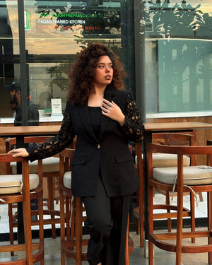 A woman with curly hair stands in a modern indoor setting with large windows and wooden bar stools, wearing a black tailored blazer with sheer black lace sleeves and black trousers.