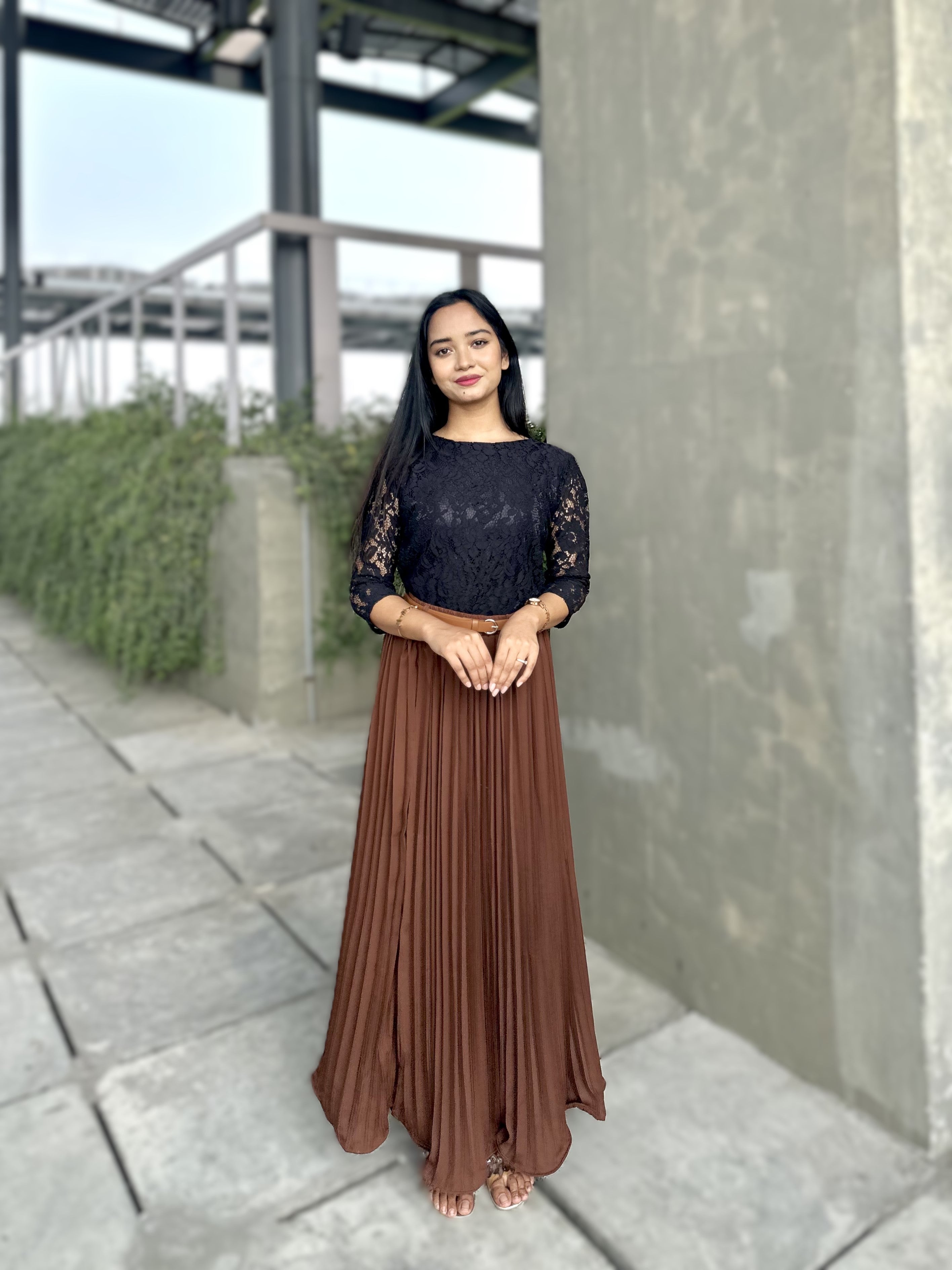 Woman wearing a black top and brown pleated skirt standing outdoors.