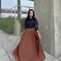 Woman wearing a black top and brown pleated skirt standing outdoors near a concrete wall.