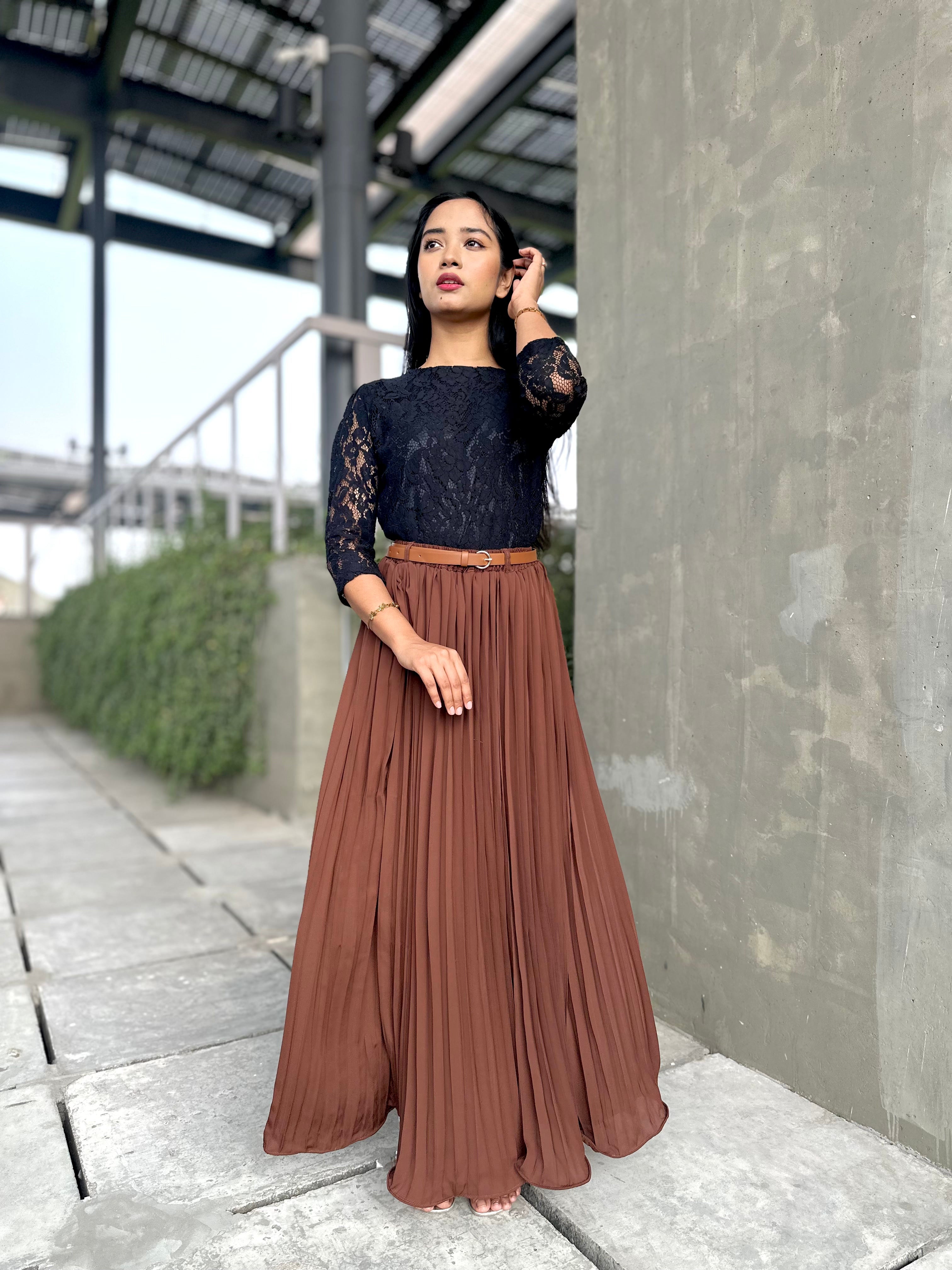 Woman wearing a black top with lace details and a brown pleated skirt standing outdoors.