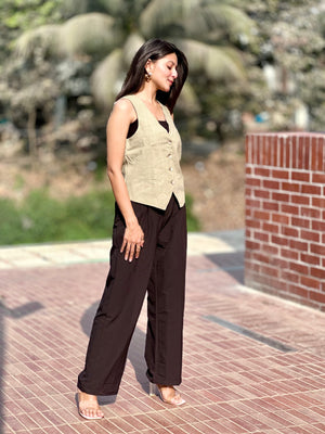 Woman in a beige vest and black pants standing on a brick path with blurred greenery in the background