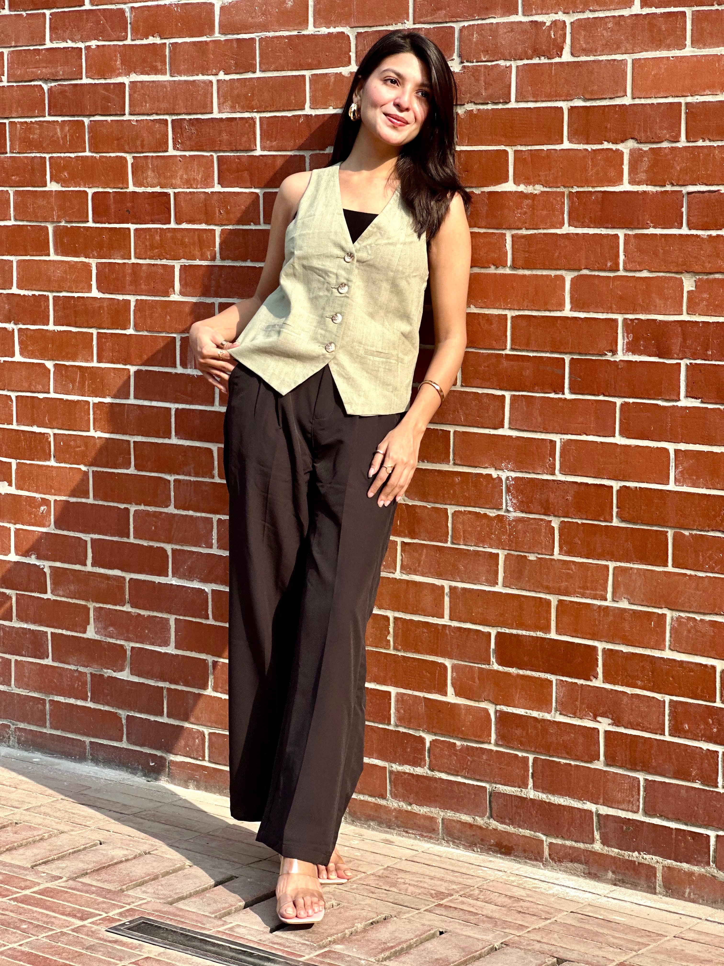 Woman in a sleeveless vest and dark pants standing against a brick wall.