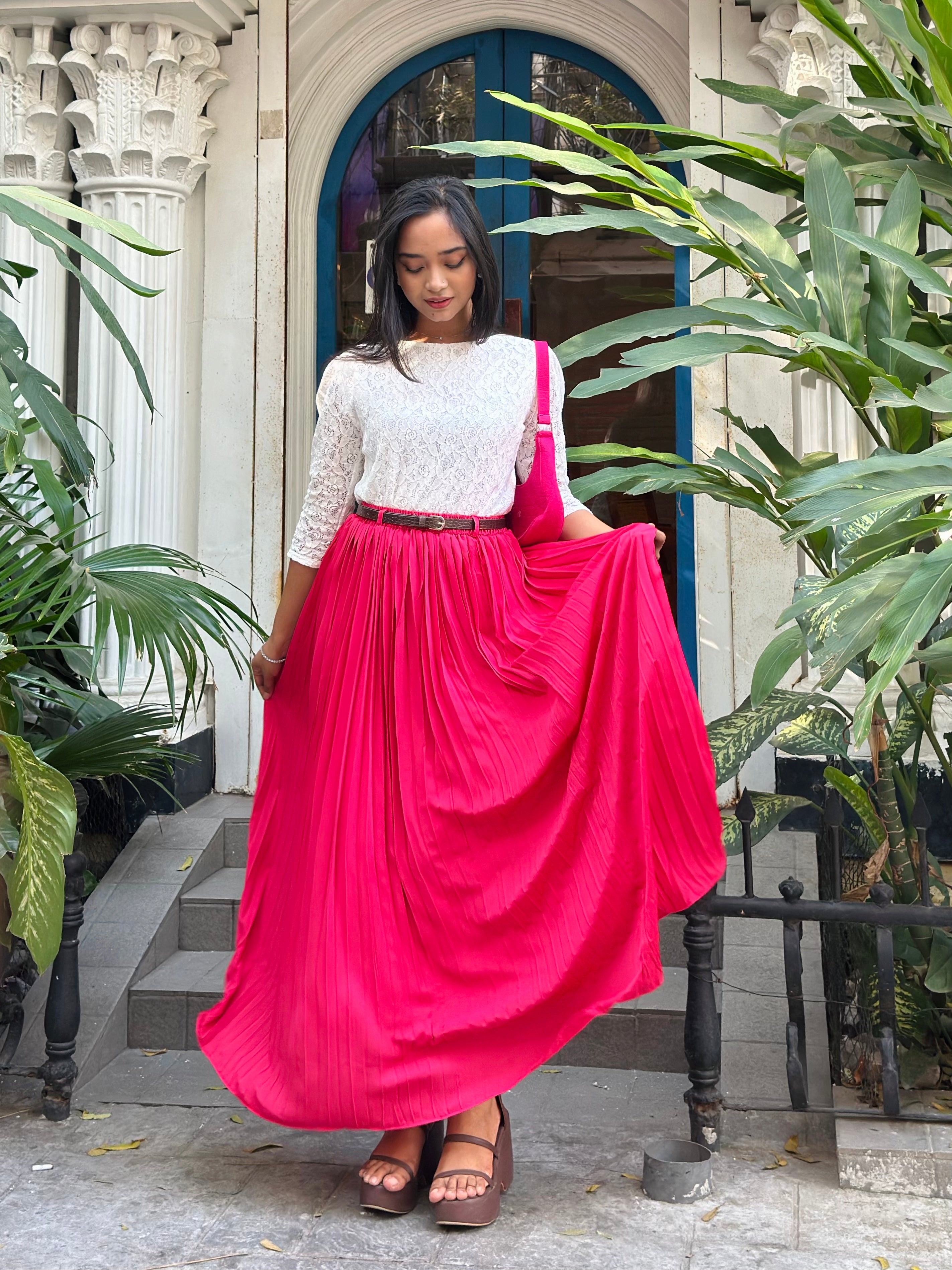 Woman in a white top and pink skirt standing outdoors with plants around.
