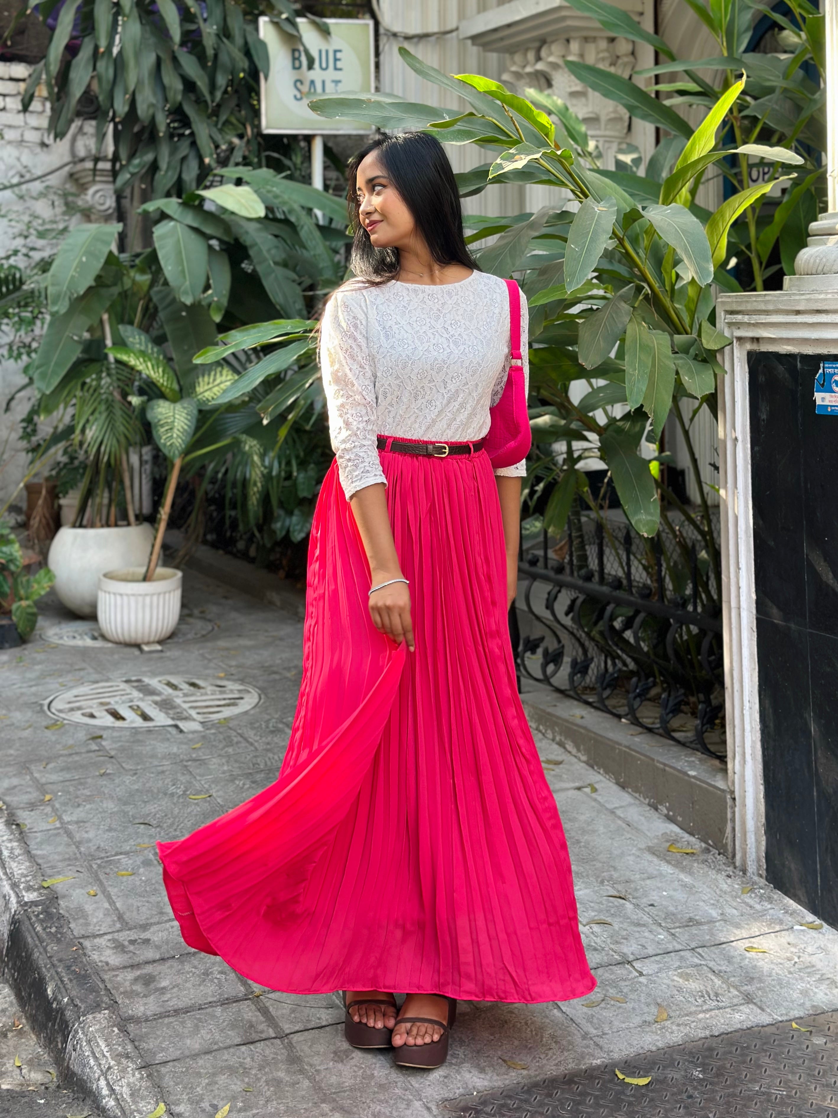 Woman in a white top and pink skirt standing outdoors with plants in the background