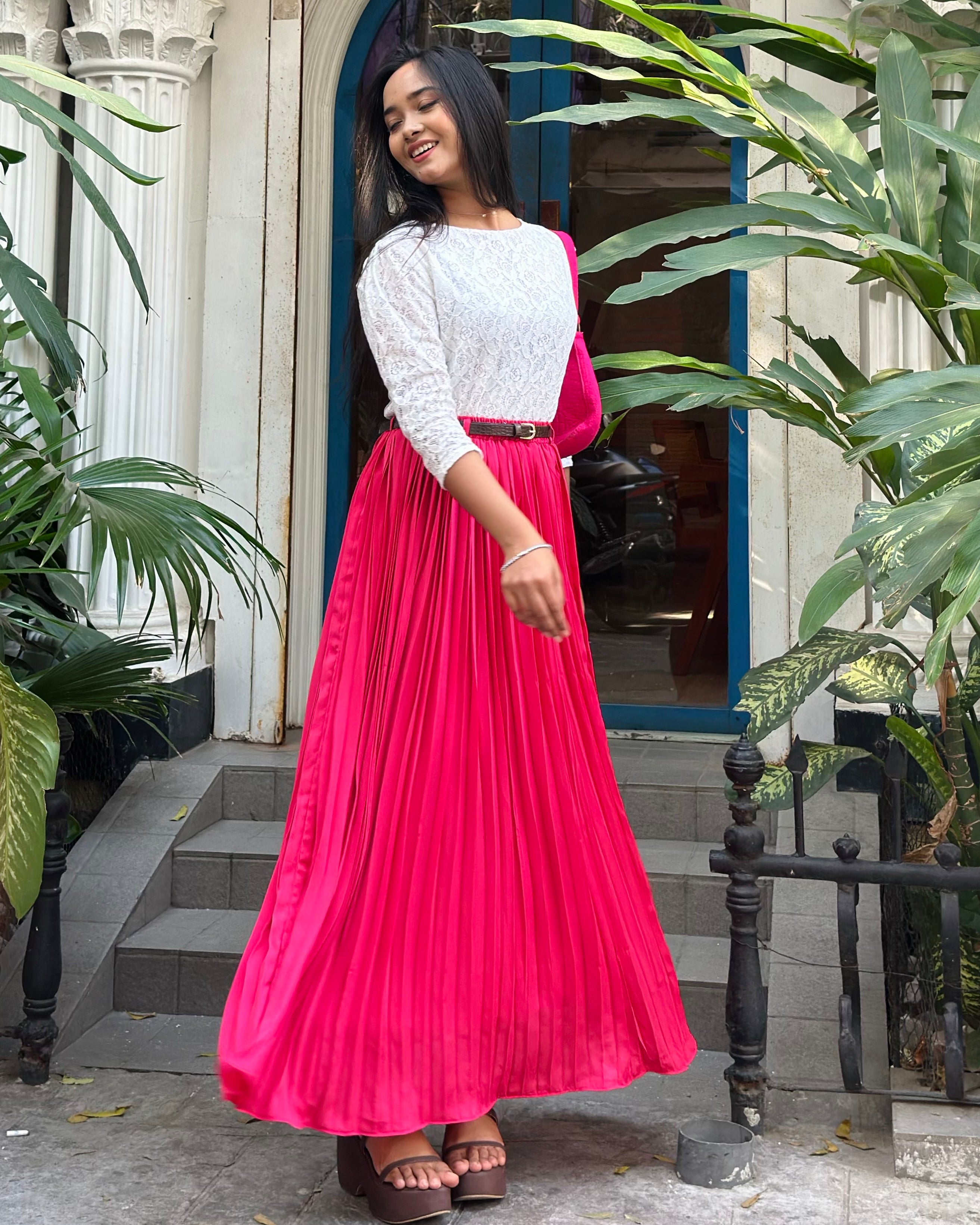 Woman in a white top and pink pleated skirt standing outdoors with plants around.