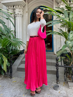 Woman in a white top and pink skirt standing outdoors with plants around.