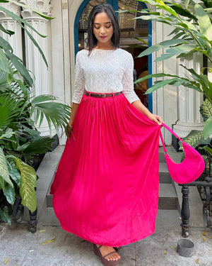 Woman holding a pink bag wearing a white top and pink skirt in an outdoor setting with plants.