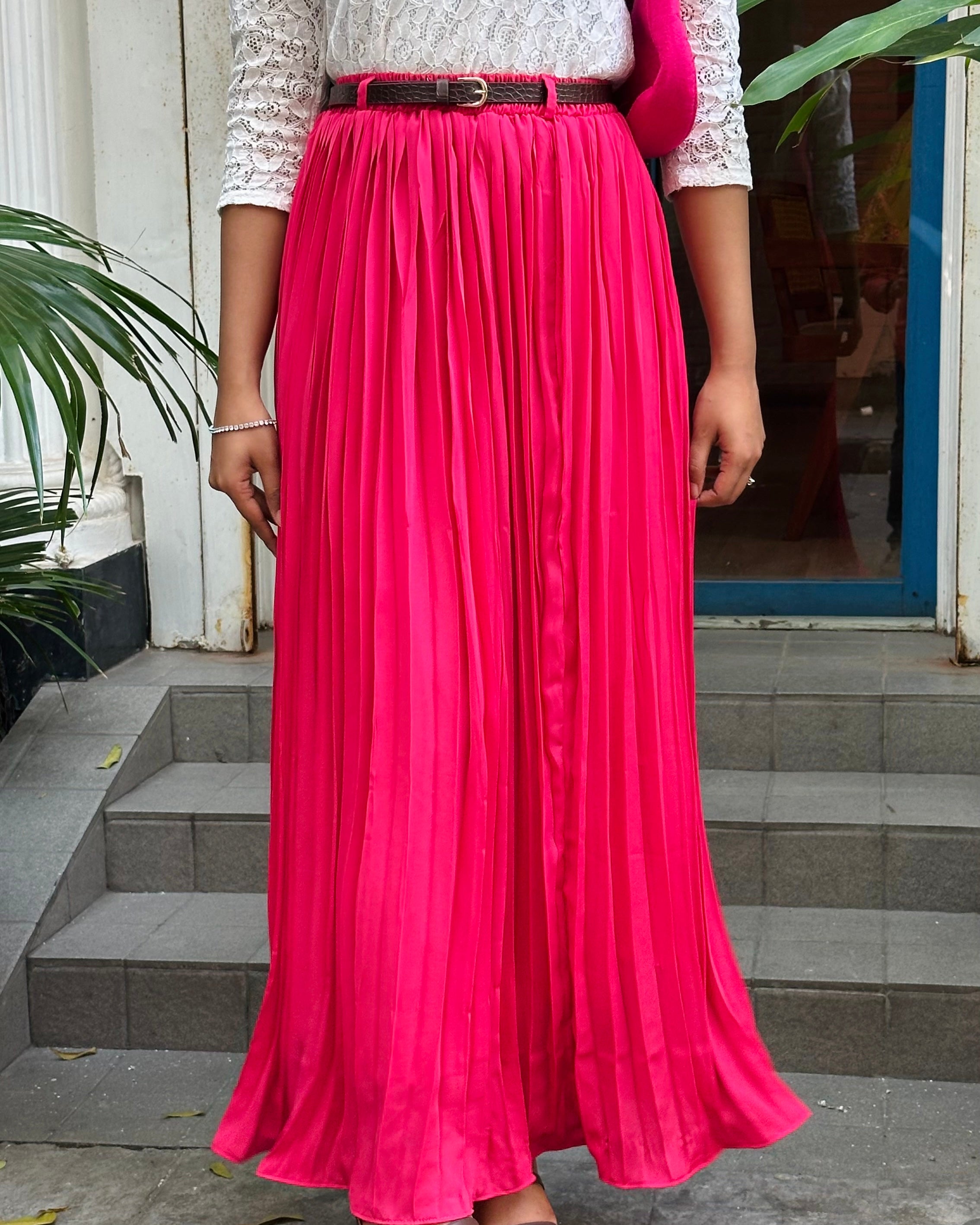 Person wearing a long pink skirt with a white top and black belt, standing on a concrete step.