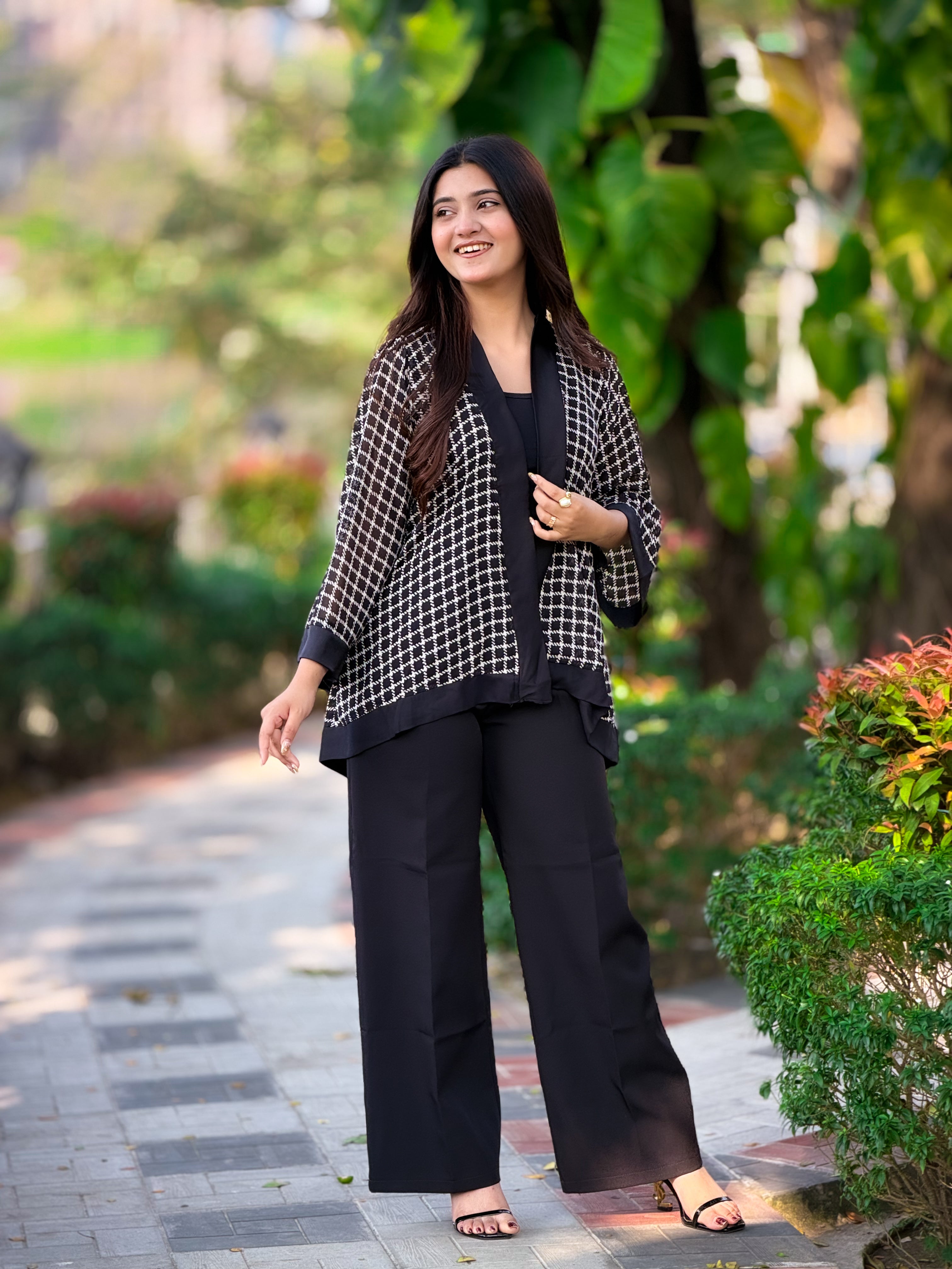 Woman in black outfit with checkered jacket standing on a pathway with greenery