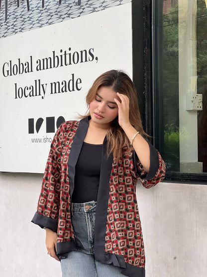Woman wearing a patterned short kimonos and jeans leaning against a wall with a sign in the background.
