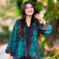 Woman wearing a teal leopard print kimono over a black outfit, standing outdoors with greenery in the background.