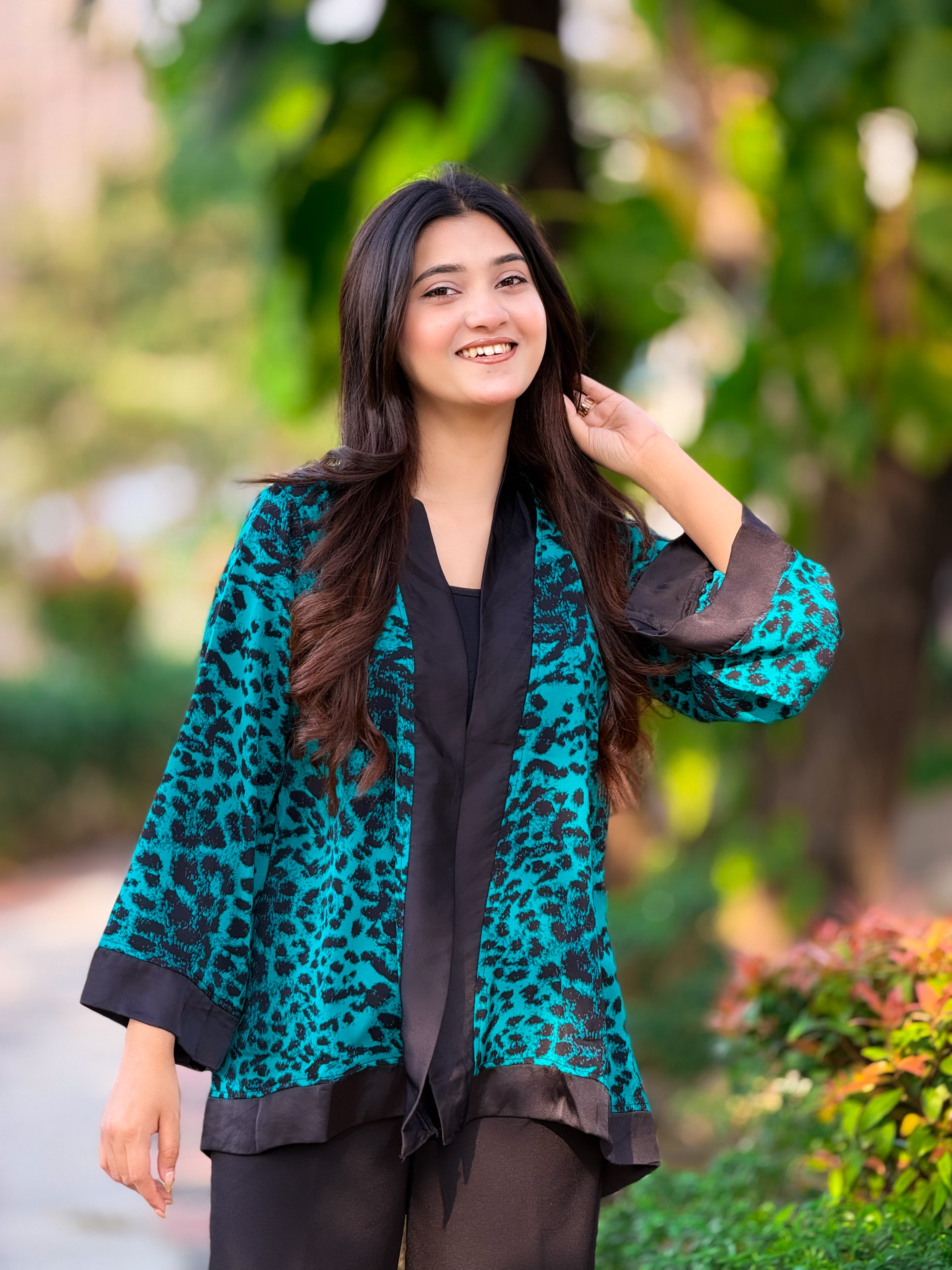 Woman wearing a teal leopard print kimono over a black outfit, standing outdoors with greenery in the background.