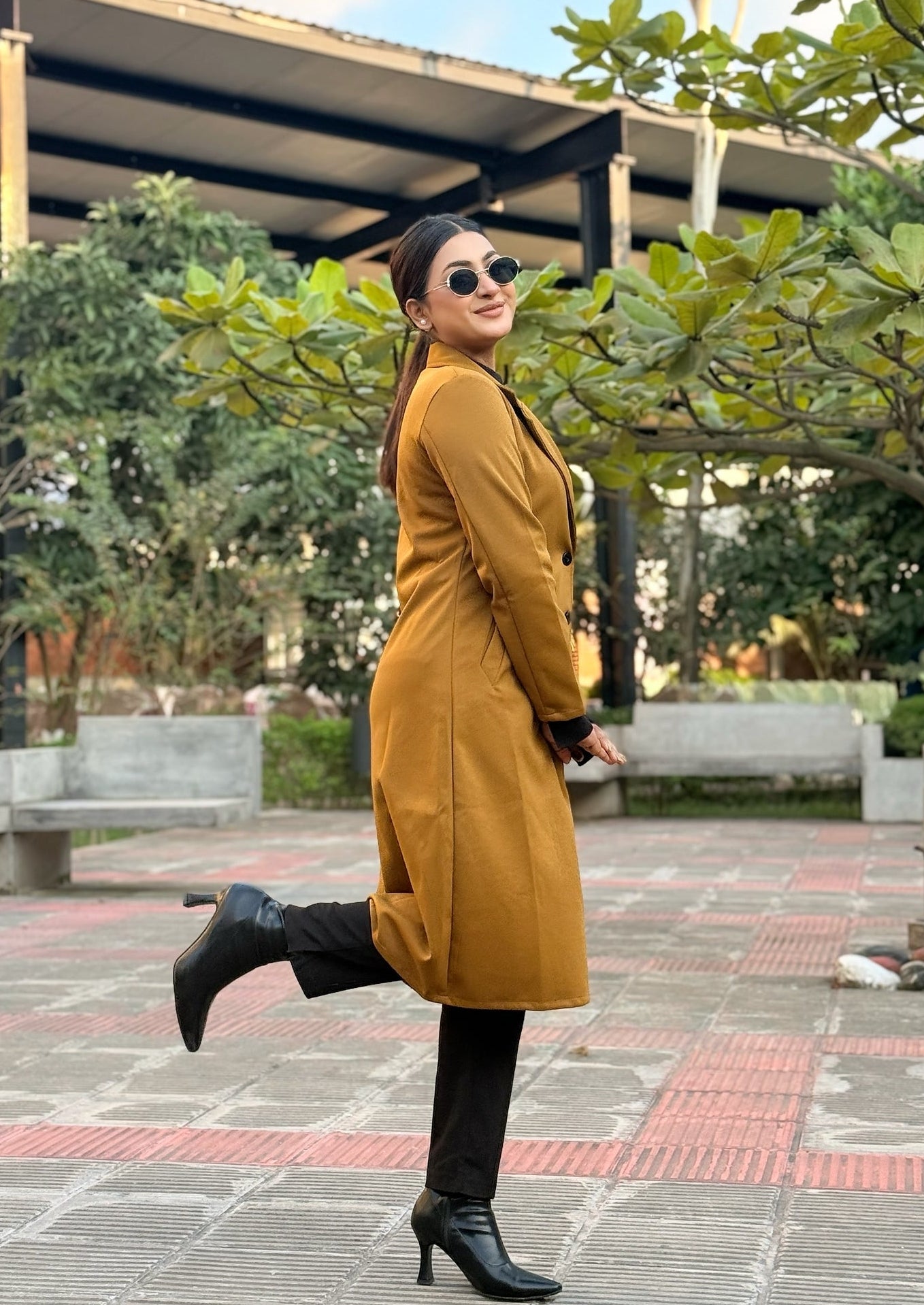 Woman in a mustard coat standing outdoors with greenery and a building in the background