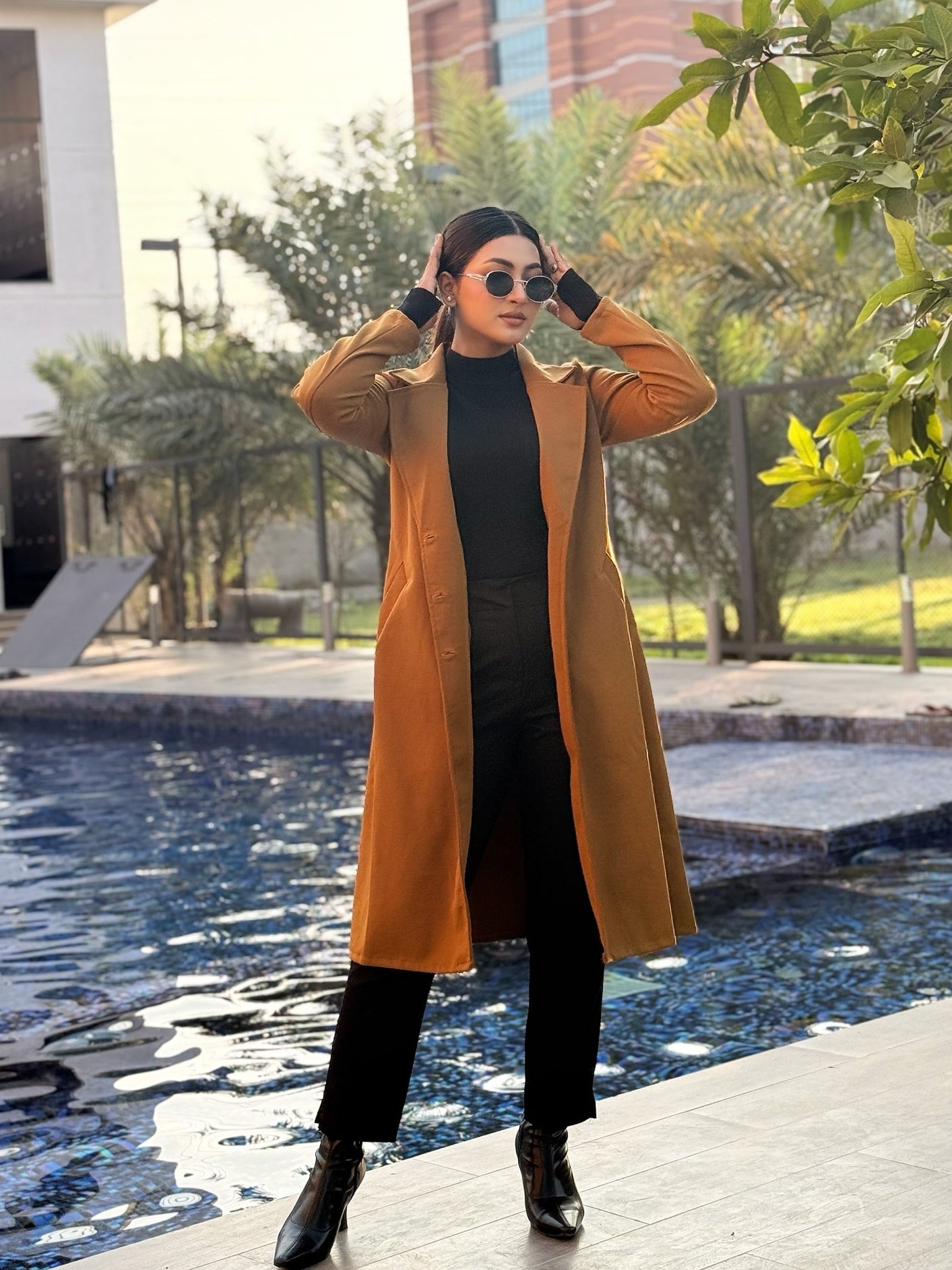 Woman in a mustard yellow coat standing by a pool with buildings and trees in the background