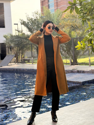 Woman in a mustard yellow coat standing by a pool with buildings and trees in the background