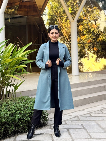 Woman in a long light blue coat standing outdoors near a modern building with greenery.
