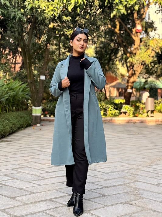 Person wearing a long light blue coat in an outdoor setting with trees and a pathway.